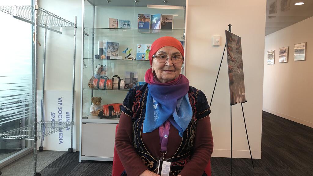 Портрет пожилой женщины, одетой в красный платок, очки и красочный шарф. Она сидит перед стеклянным экранным корпусом, содержащим книги ООН по правам человека и табличку с надписью "Создание лучшего будущего для всех".