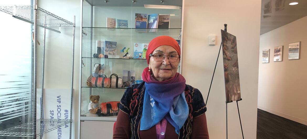 Portrait of an elderly woman wearing a red headscarf, glasses, and a colorful scarf. She is seated in front of a glass display case containing UN Human Rights books and a sign that reads 'Building a better future for all'. A UNICEF banner is visible to the left.