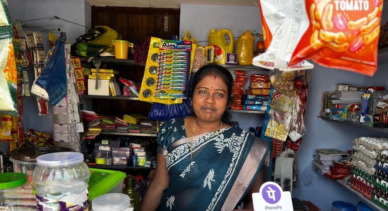 भारत में अपने छोटे, अच्छी तरह से स्टॉक किए गए सामान्य स्टोर के अंदर नीले और चांदी की साड़ी में एक भारतीय महिला का एक भारतीय महिला का चित्र भारत में अपने छोटे, अच्छी तरह से स्टॉक किए गए जनरल स्टोर के अंदर खड़ा है। दुकान विभिन्न घरेलू सामान, स्नैक्स और किराने की वस्तुओं से भरी हुई है। डिजिटल भुगतान के लिए एक PhonePe QR कोड अग्रभूमि में काउंटर पर दिखाई देता है।