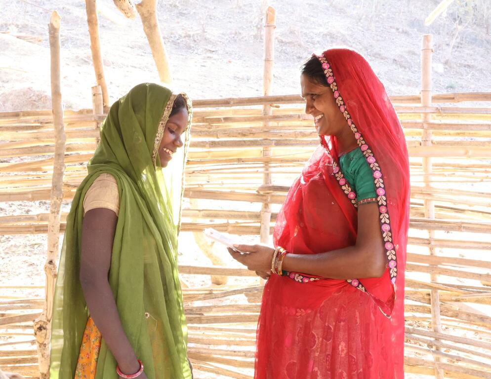 Deux femmes indiennes souriantes en saris colorés se tiennent dans un cadre rural avec une structure en bambou à l'arrière-plan. Une femme en rouge montre quelque chose sur un papier à l'autre femme en vert.