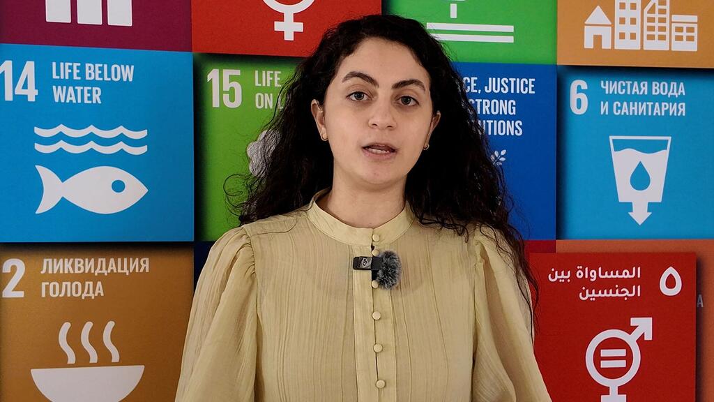 امرأة ذات شعر داكن ترتدي بلوزة بيجية وميكروفون على صدرها، تقف أمام جدار ملون يعرض أيقونات أهداف التنمية المستدامة للأمم المتحدة ونصوصًا بلغات متعددة.