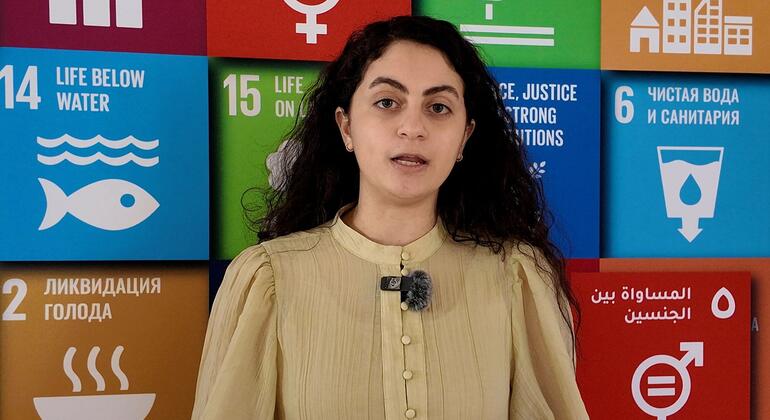 امرأة ذات شعر داكن ترتدي بلوزة بيجية وميكروفون على صدرها، تقف أمام جدار ملون يعرض أيقونات أهداف التنمية المستدامة للأمم المتحدة ونصوصًا بلغات متعددة.