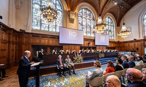 UN Secretary-General António Guterres speaking at a podium during a solemn sitting at the International Court of Justice to celebrate its 80th anniversary. Other dignitaries including King Willem-Alexander of the Netherlands, ICJ President Iwasawa Yuji, and UN officials are seated in the Great Hall of Justice.