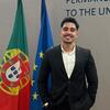 Antonio Pinho, um jovem de terno preto e camisa branca, sorri enquanto está em frente às bandeiras das Nações Unidas, de Portugal e da União Europeia, na Missão Permanente junto às Nações Unidas.