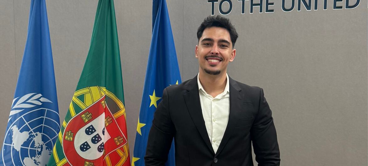 Antonio Pinho, um jovem de terno preto e camisa branca, sorri enquanto está em frente às bandeiras das Nações Unidas, de Portugal e da União Europeia, na Missão Permanente junto às Nações Unidas.