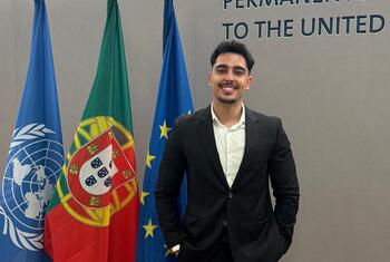 Antonio Pinho, um jovem de terno preto e camisa branca, sorri enquanto está em frente às bandeiras das Nações Unidas, de Portugal e da União Europeia, na Missão Permanente junto às Nações Unidas.