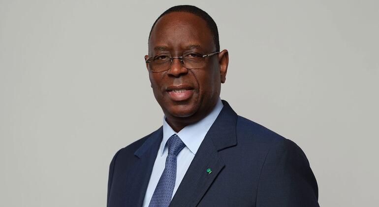 Профессиональное фото политика из Африки Салла в тёмном костюме, светло-голубой рубашке, галстуке с узором и очках. На его лацкане зелёная булавка, он слегка улыбается на однотонном сером фоне.