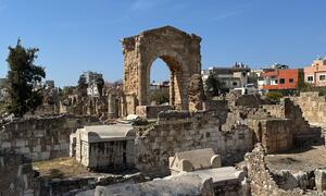 Ancient stone ruins of the historical city of Tyre in Lebanon, featuring a prominent stone archway and sarcophagi under a clear blue sky, with modern buildings visible in the background.