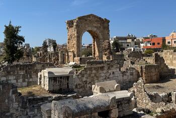 Ancient stone ruins of the historical city of Tyre in Lebanon, featuring a prominent stone archway and sarcophagi under a clear blue sky, with modern buildings visible in the background.