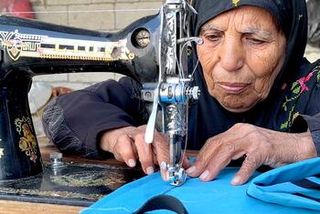 امرأة مسنة من غزة تدعى نغين بن عيسى، ترتدي حجابا أسود، تركز على تشغيل ماكينة خياطة سوداء قديمة لإصلاح قطعة ملابس زرقاء. هذا العمل ضرورة لها بعد أن قطعت الحرب المساعدات الاجتماعية عن عائلتها.