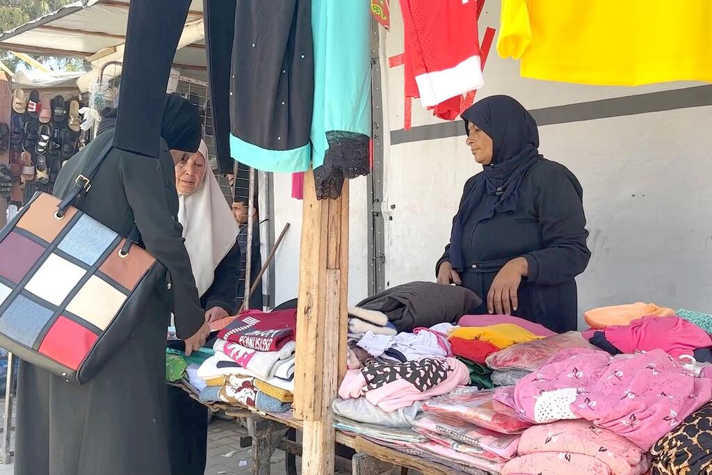 جود كواريا، امرأة فلسطينية وأم، تدير كشكاً صغيراً لبيع الملابس في الهواء الطلق بالقرب من مستشفى ناصر في غزة. تظهر الصورة توازنها بين عملها في بيع الملابس ومسؤولياتها العائلية في بيئة تفتقر إلى الاحتياجات الأساسية.