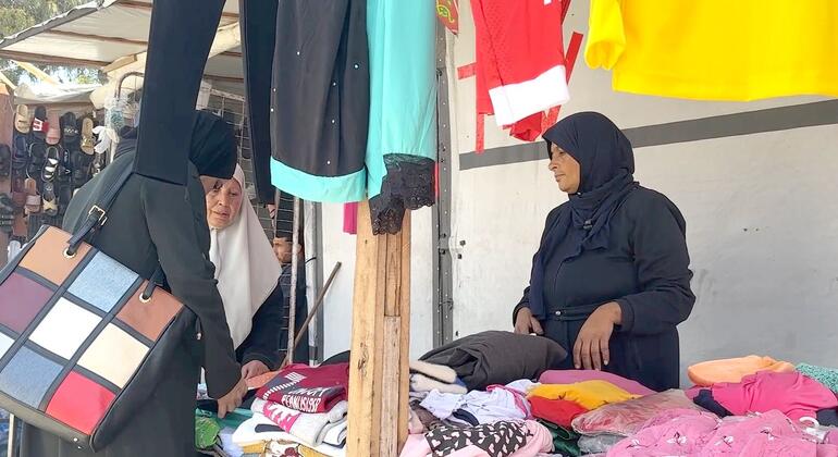 جود كواريا، امرأة فلسطينية وأم، تدير كشكاً صغيراً لبيع الملابس في الهواء الطلق بالقرب من مستشفى ناصر في غزة. تظهر الصورة توازنها بين عملها في بيع الملابس ومسؤولياتها العائلية في بيئة تفتقر إلى الاحتياجات الأساسية.