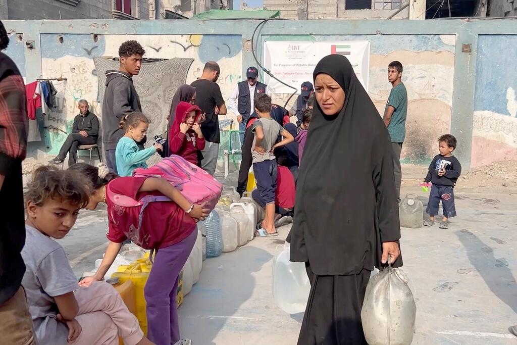 امرأة ترتدي حجابًا أسود وأطفال ينتظرون في طابور لتلقي المياه من حاويات كبيرة في مخيم للنازحين في غزة.