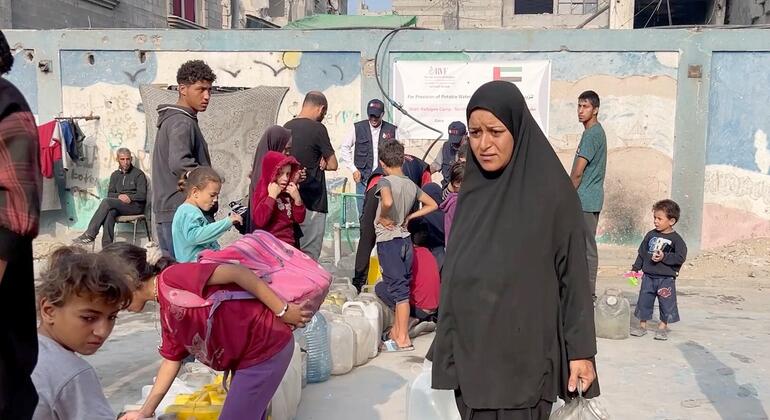 امرأة ترتدي حجابًا أسود وأطفال ينتظرون في طابور لتلقي المياه من حاويات كبيرة في مخيم للنازحين في غزة.