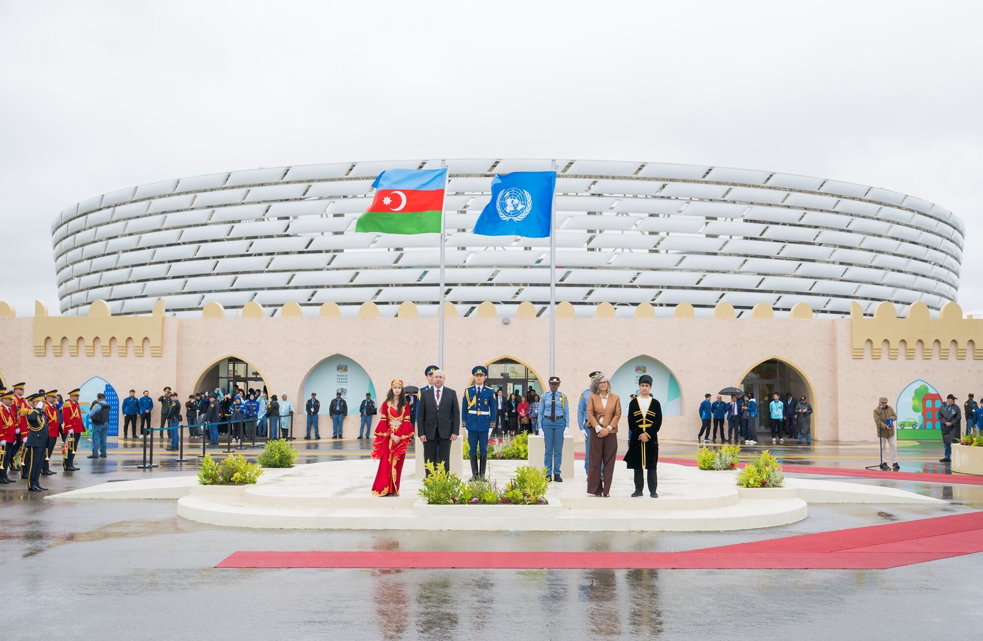 باکو میں ورلڈ اربن فورم 13 کے دوران ایک بیرونی تقریب کے موقع پر معزز شخصیات اور عہدیدار ایک منچ پر کھڑے ہیں، جبکہ ایک بڑی عمارت کے سامنے اقوام متحدہ اور آذربائیجان کے جھنڈے لہرا رہے ہیں۔