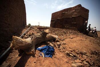 Firewood used to dry bricks in a massive oven in El Fasher, North Darfur, Sudan. The image highlights the environmental impact of brick production, showing a large pile of dirt and broken bricks with a tree stump in the foreground.