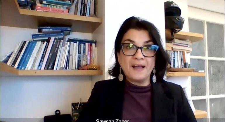 Header photo of Sawsan Zaher during an interview, wearing glasses and a black jacket over a purple bow. She is sitting in front of library shelves full of books.