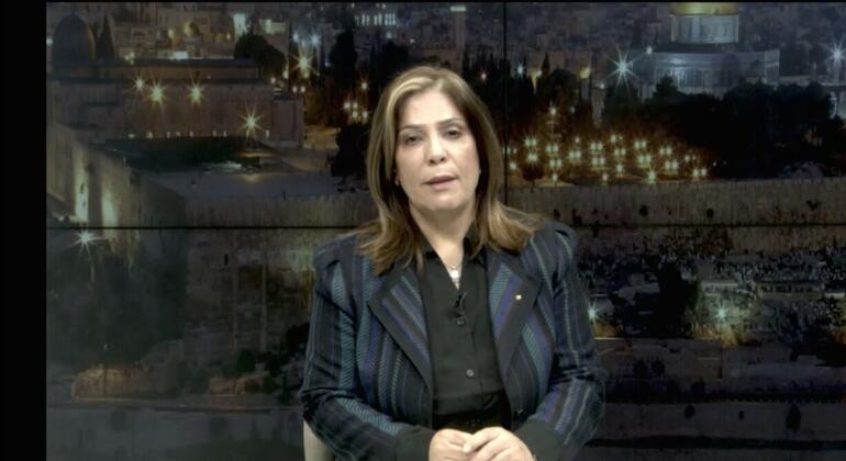 قناة أخبار نسائية ذات شعر بني تجلس في استوديو مع خلفية تظهر منظر مضيء على القدس في الليل، بما في ذلك قبة الصخرة.