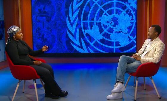Mwanamke na mwanamume wanakaa kwenye viti vikubwa vikubwa vikubwa vinavyoangaliana wakati wa mahojiano katika studio moja wakiwa na nembo kubwa ya Umoja wa Mataifa kwenye skrini ya bluu nyuma yao.