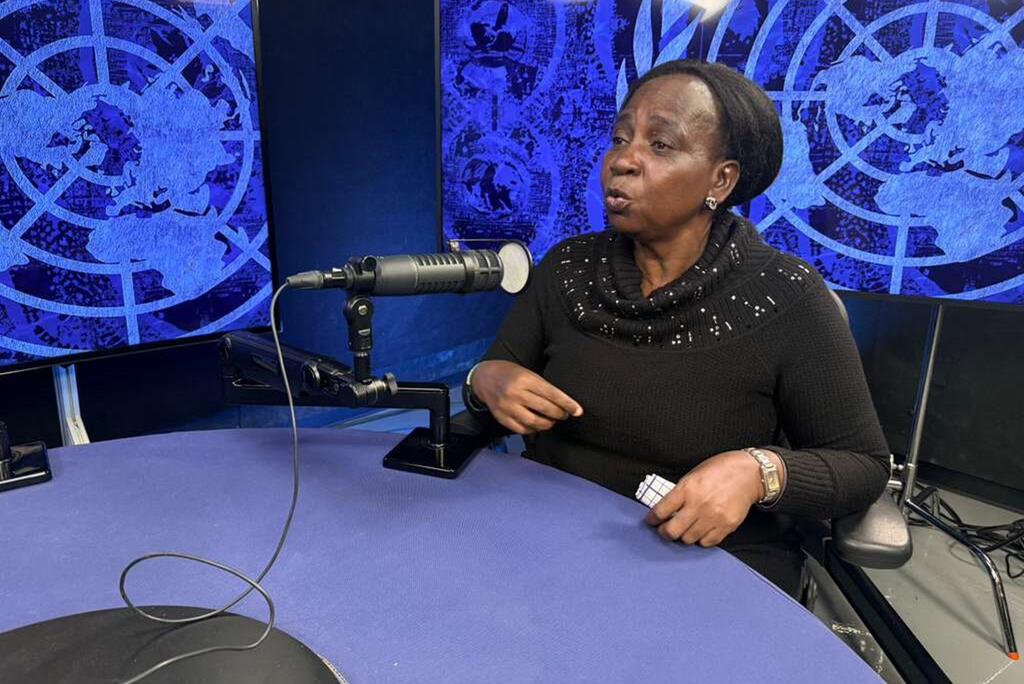 Mwanamke mmoja Mwafrika mwenye kifuniko kichwani ameketi kwenye dawati katika studio, akizungumza kwenye kipaza sauti. Nyuma yake kuna skrini za bluu zinazoonyesha ramani ya ulimwengu.