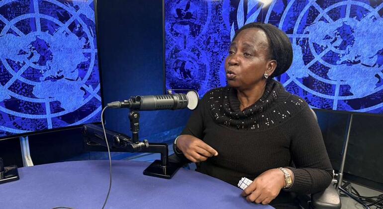 Mwanamke mmoja Mwafrika mwenye kifuniko kichwani ameketi kwenye dawati katika studio, akizungumza kwenye kipaza sauti. Nyuma yake kuna skrini za bluu zinazoonyesha ramani ya ulimwengu.