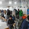 Группа афганских беженцев и мигрантов, включая мужчин и мальчиков, с багажом в большом зале временного приюта, некоторые сидят на деревянных скамейках, другие стоят.