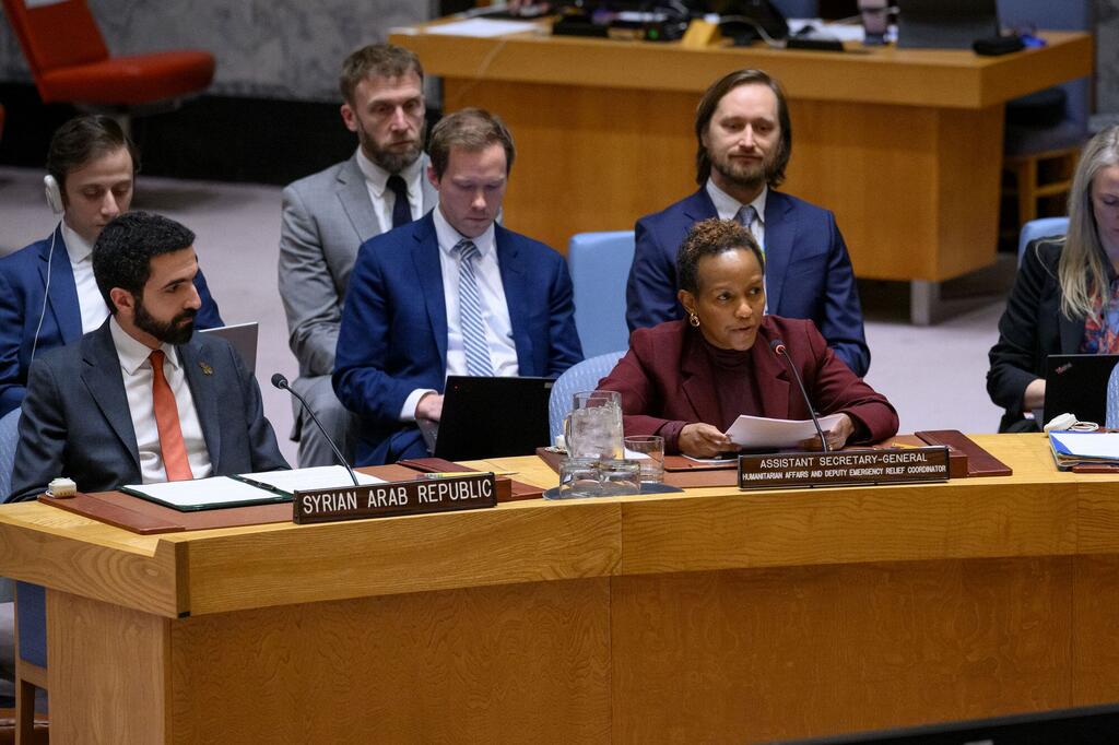 Une femme en blazer maroon s'exprimant à une table de conférence avec une plaque portant le nom de " secrétaire général adjoint aux affaires humanitaires et coordonnateur adjoint des secours d'urgence ". Derrière elle, un homme est assis à une table avec une plaque portant le nom de "République arabe syrienne".