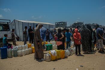Families in Gaza gather around a water truck in a displacement camp, collecting water in jerrycans. In the background are damaged buildings and tents. This photo illustrates the severe water insecurity affecting families in the Gaza Strip.