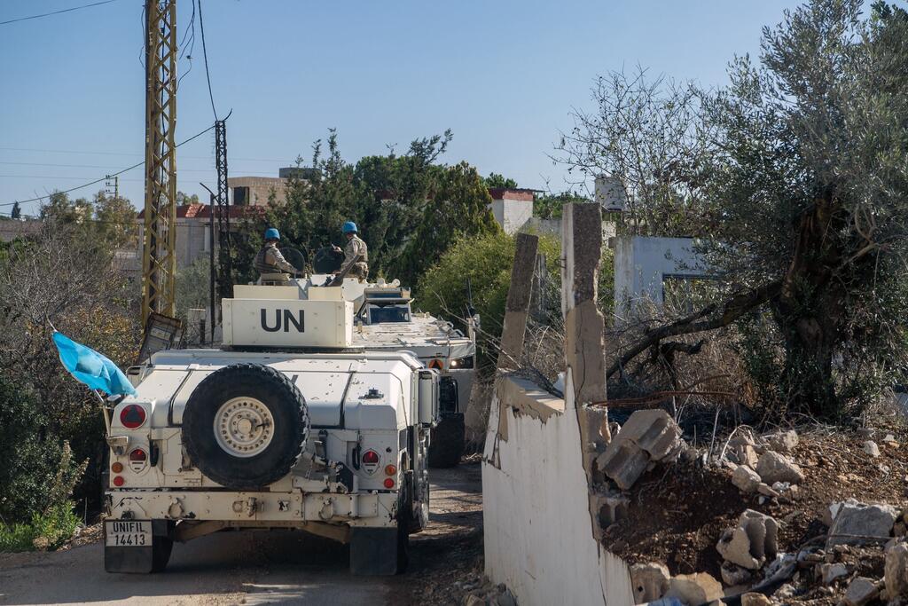 Des véhicules de maintien de la paix de la FINUL, avec des casques bleus, patrouillent sur une route au Liban, en passant devant un mur de béton endommagé et des oliviers.