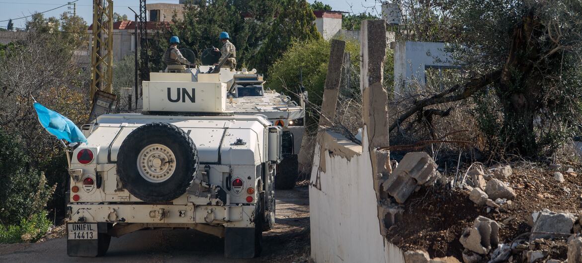 Veículos de manutenção da paz da UNIFIL, com capacetes azuis, patrulham uma estrada no Líbano, passando por um muro de concreto danificado e oliveiras.