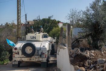 UNIFIL peacekeeping vehicles with blue helmets patrol a road in Lebanon, passing by a damaged concrete wall and olive trees.