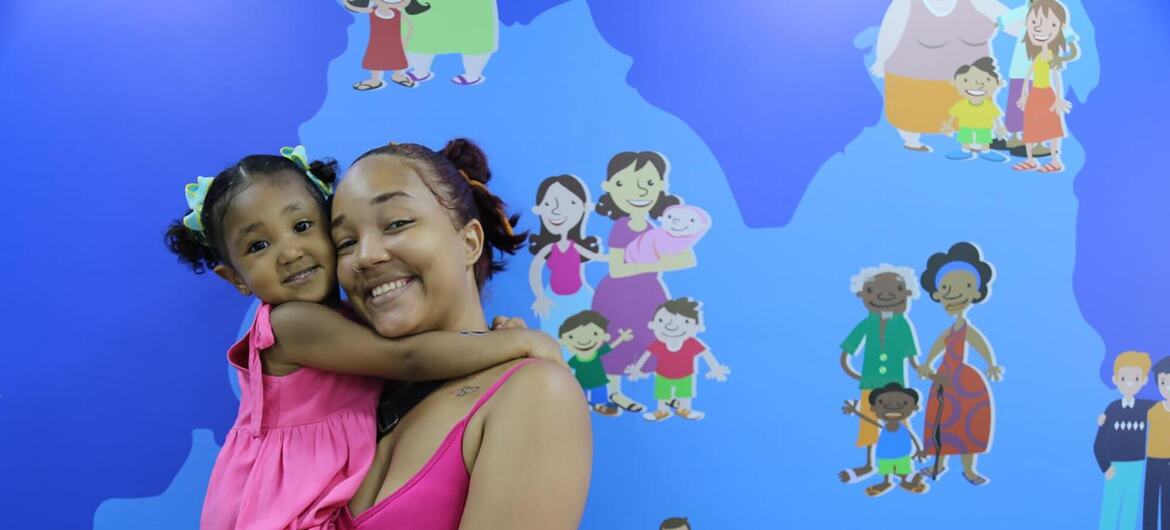 Uma mulher sorridente de vestido rosa abraça uma menina diante de uma parede azul com ilustrações de desenhos animados de várias famílias.