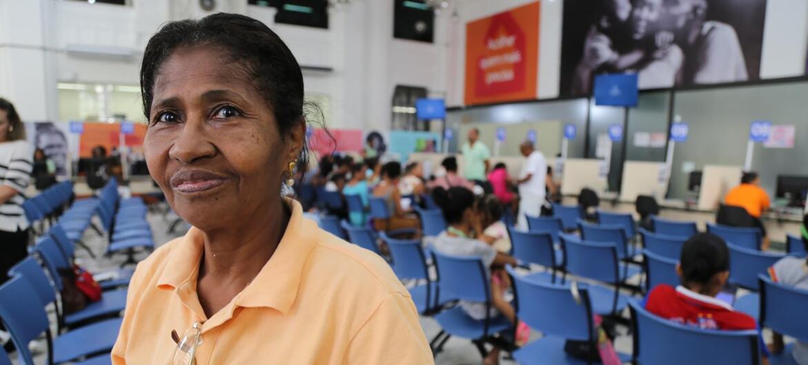 Uma mulher de poloshirt laranja sorri para a câmera enquanto está sentada em uma sala de espera cheia de cadeiras azuis e outras pessoas.
