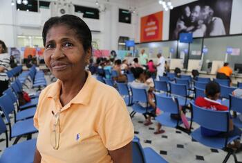 Uma mulher de poloshirt laranja sorri para a câmera enquanto está sentada em uma sala de espera cheia de cadeiras azuis e outras pessoas.