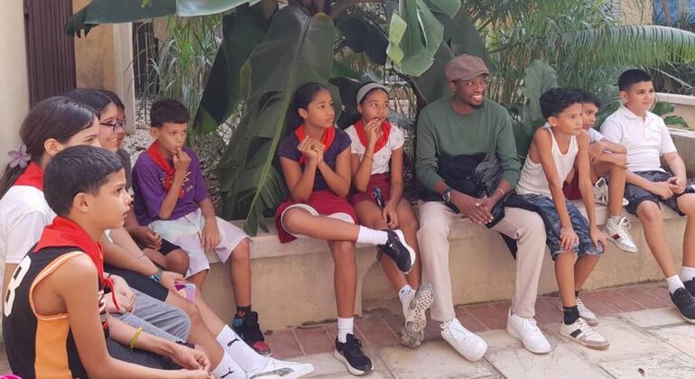 Kikundi cha wanafunzi wadogo na mtu mzima wameketi pamoja kwenye benchi la jiwe huko Havana, Cuba, wakifanya shughuli za kujifunza zinazohusiana na elimu ya lugha ya Kiswahili.