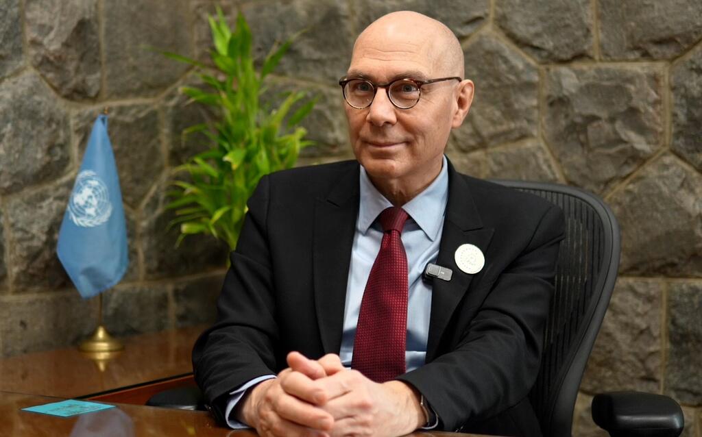 Um retrato formal de Volker Türk, um homem careca usando óculos, um terno escuro e uma gravata vermelha, sentado em uma mesa com uma bandeira da ONU no fundo.