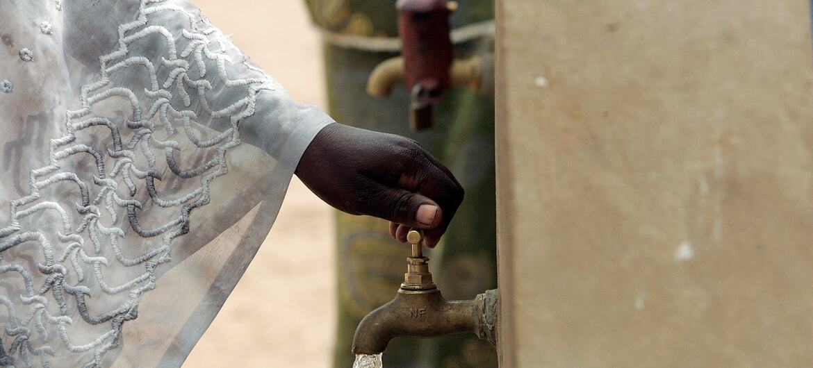 A close-up of a woman's hand turning on a communal water faucet in Ndombil, Senegal, with clean water flowing out.