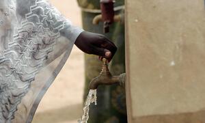 A close-up of a woman's hand turning on a communal water faucet in Ndombil, Senegal, with clean water flowing out.