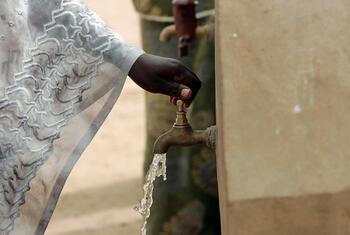 A close-up of a woman's hand turning on a communal water faucet in Ndombil, Senegal, with clean water flowing out.