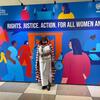 Mwanamke katika mavazi ya kitamaduni ya Maasai amesimama mbele ya bendera kubwa ya UN Women kwa CSW70, ambayo inasema 'Haki. Haki. Hatua. Kwa Wanawake na Wasichana Wote'.