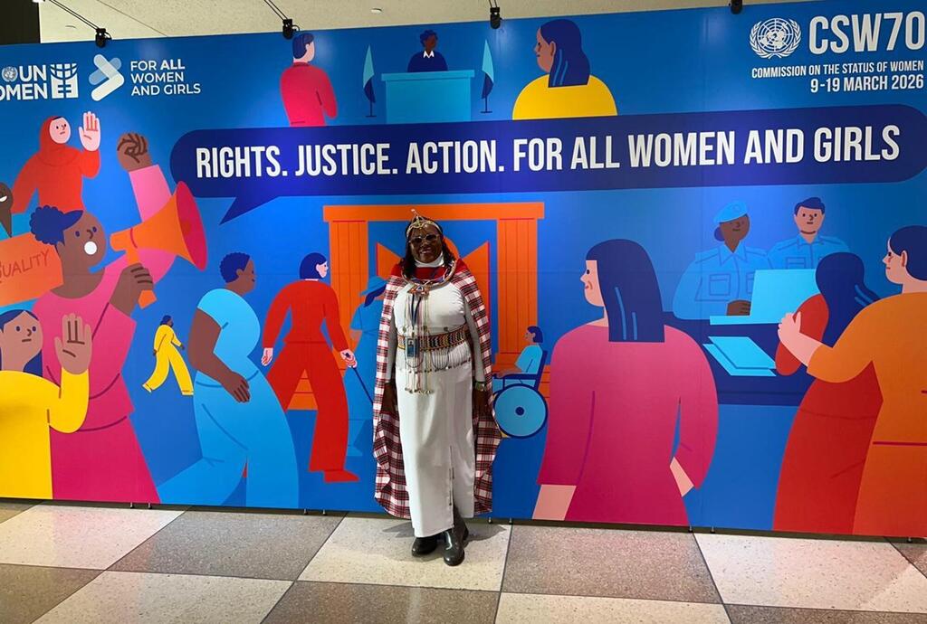 Mwanamke katika mavazi ya kitamaduni ya Maasai amesimama mbele ya bendera kubwa ya UN Women kwa CSW70, ambayo inasema 'Haki. Haki. Hatua. Kwa Wanawake na Wasichana Wote'.