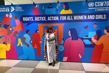 Mwanamke katika mavazi ya kitamaduni ya Maasai amesimama mbele ya bendera kubwa ya UN Women kwa CSW70, ambayo inasema 'Haki. Haki. Hatua. Kwa Wanawake na Wasichana Wote'.