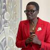 Mwanamke mweusi amevaa blazer nyekundu, miwani, na mkufu wa lulu akizungumza.