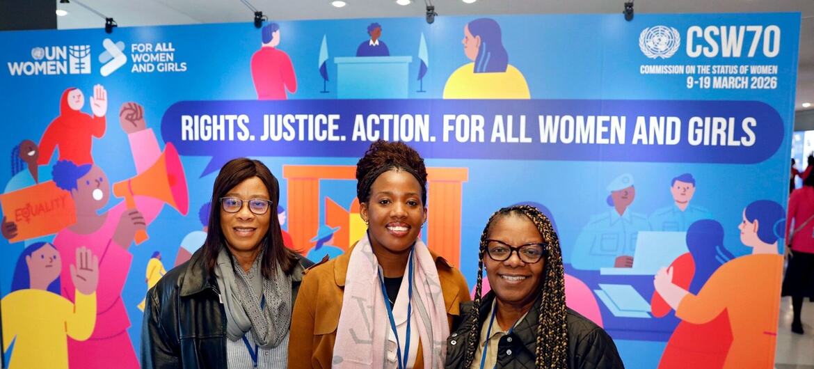 Três mulheres negras sorridentes posando para uma foto na frente de uma bandeira azul da ONU Mulheres CSW70.