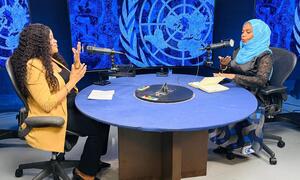 Wanawake wawili wameketi kwenye meza ya mviringo yenye rangi ya bluu wakiwa na maikrofoni, wakirekodi podcast katika studio yenye alama ya Umoja wa Mataifa.
