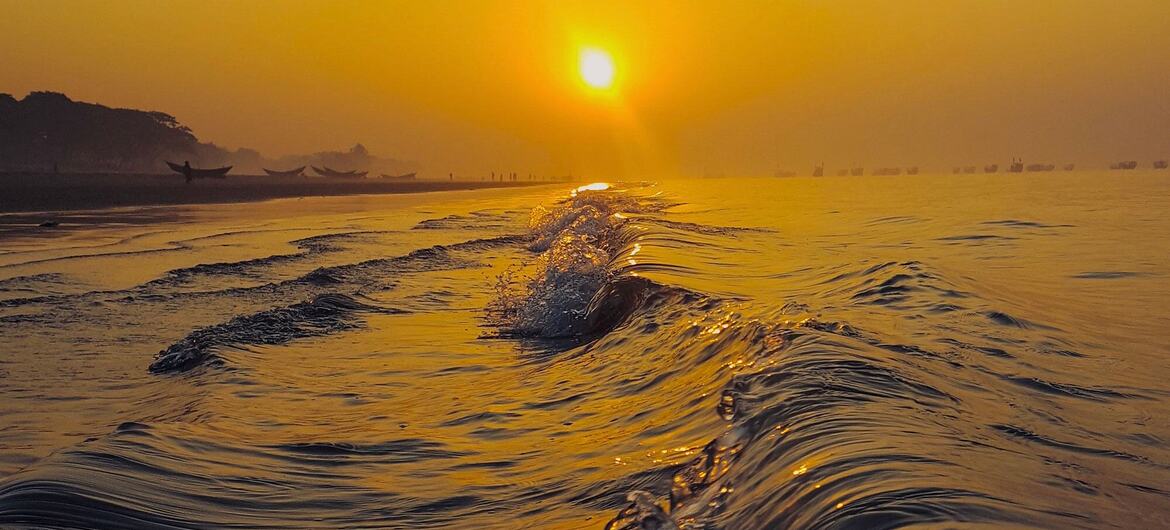 Waves surge on the beach at Kuakata, Bangladesh during a golden sunset, with the sun low on the horizon and boats visible in the distance.