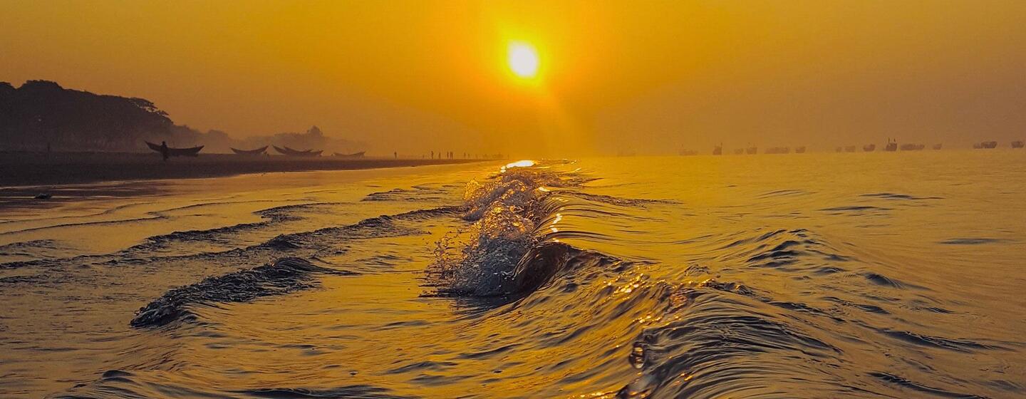 Les vagues déferlent sur la plage de Kuakata, au Bangladesh, pendant un coucher de soleil doré, avec le soleil bas à l'horizon et des bateaux visibles au loin.