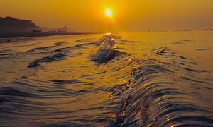 Waves surge on the beach at Kuakata, Bangladesh during a golden sunset, with the sun low on the horizon and boats visible in the distance.