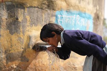 Sara, uma jovem estudante indiana de uniforme azul, bebe água de uma torneira metálica do lado de fora de sua escola na aldeia de Adol, no estado de Rajasthan. A torneira faz parte de um sistema de água resistente ao clima que fornece água potável limpa à comunidade.