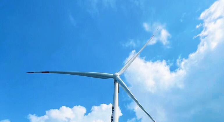 孟子晓站在一艘船的甲板上，身穿橙色救生衣并佩戴太阳镜，背景是蓝天白云，远处可见一座大型海上风力发电机。